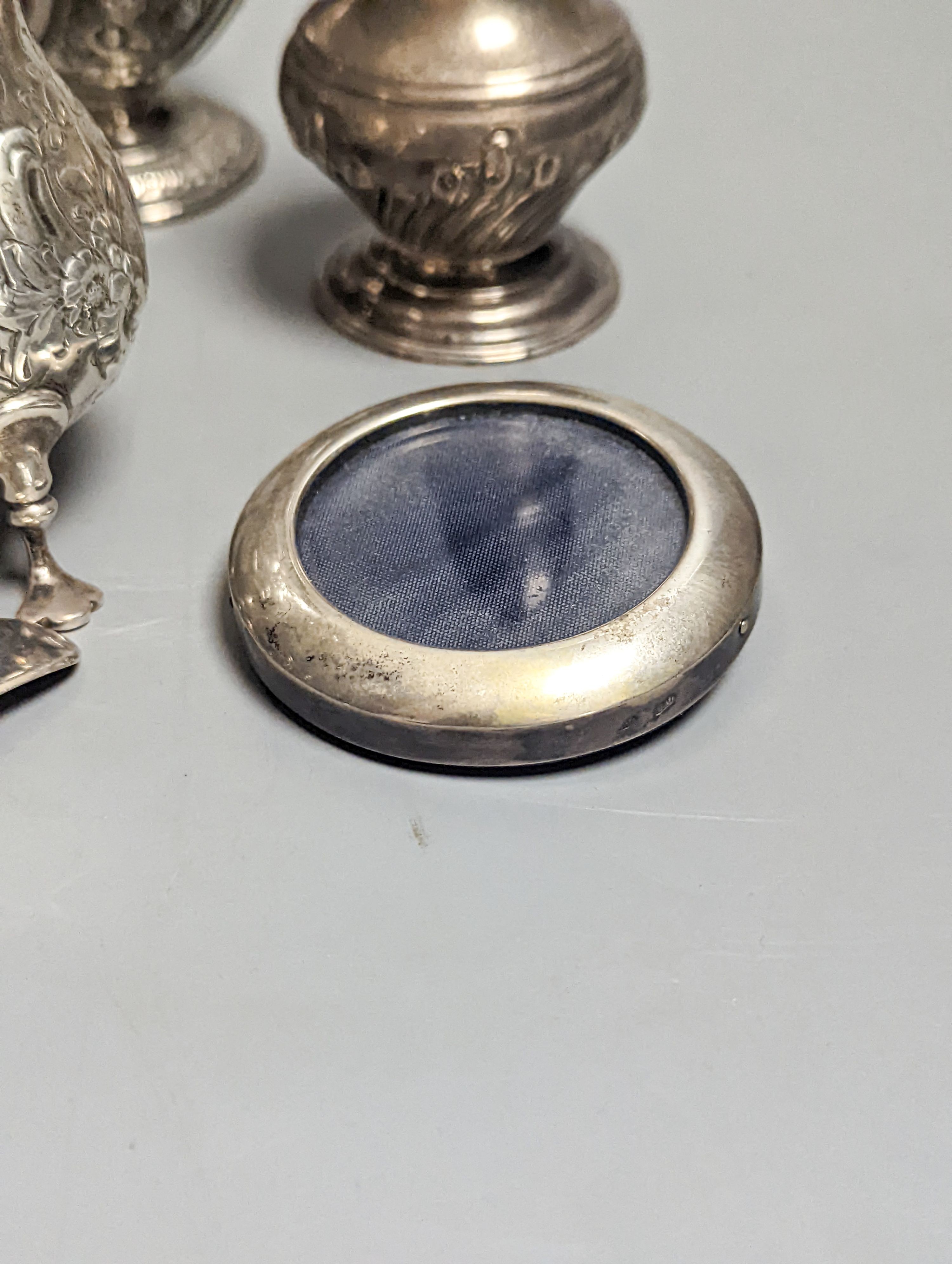 A Victorian embossed silver cream jug, London, 1883, 92mm, two 18th century silver pepperettes, one with plated cover, four later condiments (two silver), a silver spoon and small 800 standard photograph frame.